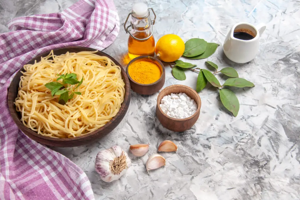 Wooden bowl of spaghetti pasta without sauce, with herb garnish in centre. Surrounded by ingredients, including garlic, salt, spices, oil, lemon | Image for: Can you freeze cooked pasta without sauce?